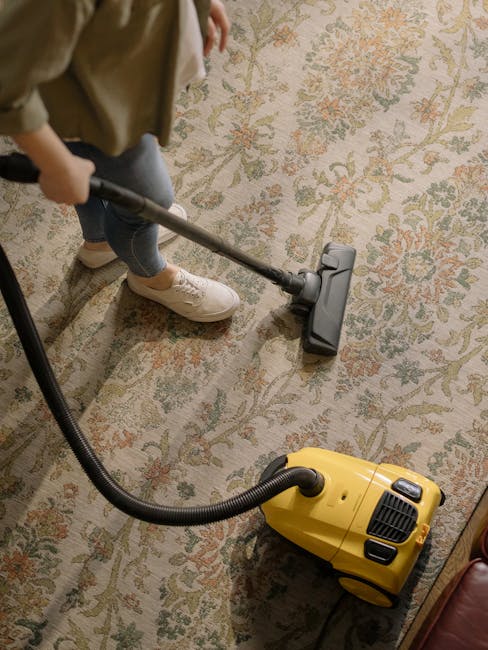 A person vacuuming a decorative patterned carpet in a residential or commercial setting. The individual, wearing casual shoes and jeans, is operating a yellow and black vacuum cleaner with a long hose attachment. The carpet features floral motifs in muted tones, and the area is well-lit, highlighting its clean and maintained condition. The image emphasizes surface cleaning and deep cleaning processes, indicative of routine domestic or commercial sanitisation. Carpet Cleaners Catford specializes in professional cleaning services across residential and business premises, ensuring thorough hygiene and surface care.