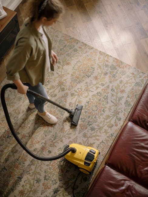 A woman is vacuuming a patterned area rug with a yellow canister vacuum cleaner in a living room. The rug features a floral design in soft pastel colors and is placed on a wooden floor. The woman is dressed in casual clothing, wearing a beige jacket, blue jeans, and sneakers. The room has natural lighting, and part of a red leather sofa is visible on the right side of the image. This scene illustrates surface cleaning performed as part of domestic cleaning services, highlighting the importance of maintaining hygiene and cleanliness in residential spaces. Carpet Cleaners Catford specializes in deep cleaning and sanitisation to ensure a hygienic environment.