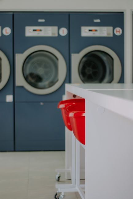 The image depicts a commercial laundry room with two front-loading washing machines in blue, positioned side by side against a light-colored wall. The machines appear clean and well-maintained, with digital display panels and controls visible on the top front sections. In the foreground, there is a white countertop with a smooth surface, and several red plastic laundry baskets neatly stacked on a white mobile cart with small wheels. The room is well-lit, showcasing a tidy and hygienic environment suited for surface cleaning and sanitisation, appropriate for a professional cleaning service like Carpet Cleaners Catford – SE6 emergency cleaning same day service in Catford. The area appears organized to facilitate efficient laundry processing and deep cleaning procedures, emphasizing hygiene and upkeep in a commercial setting.