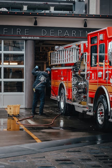 A firefighter in uniform is cleaning the exterior of a red fire truck with a hose outside a fire department building. The scene shows wet concrete pavement beneath the truck, which is parked in front of a glass entrance with a sign reading 'FIRE DEPARTMENT.' The building features a modern architectural style with large glass windows and metal framing, and the lighting suggests an overcast or early evening setting. The firefighter appears to be performing surface cleaning or sanitisation of the vehicle, ensuring it is thoroughly clean and ready for emergency response. The scene emphasizes professional equipment and meticulous cleaning processes typical of emergency service vehicles, consistent with the highly visible structure and maintenance of firefighting assets. Carpet Cleaners Catford specializes in surface cleaning and sanitisation services, ensuring hygiene and readiness of emergency and commercial equipment.